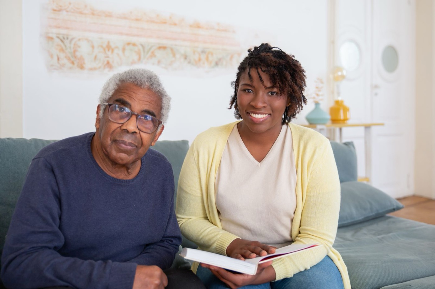 A Goldenview direct support professional sitting with an older man on his living room sofa, reading together