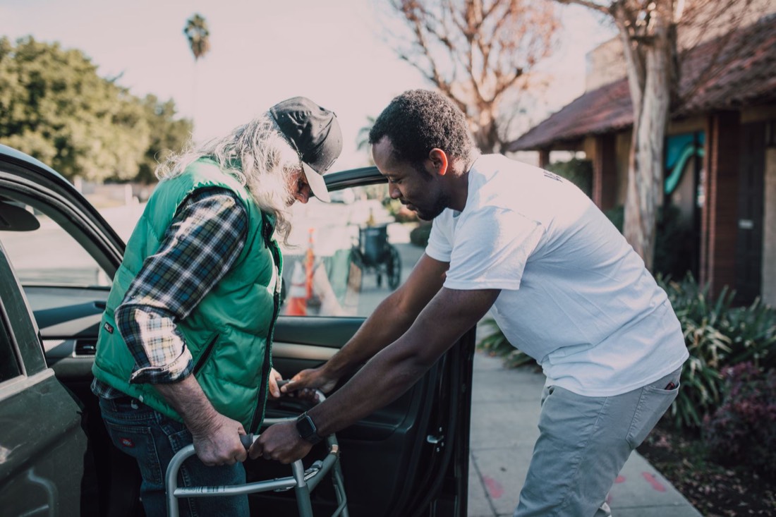 A caregiver helping an older man with a walker step out of a car outside his home